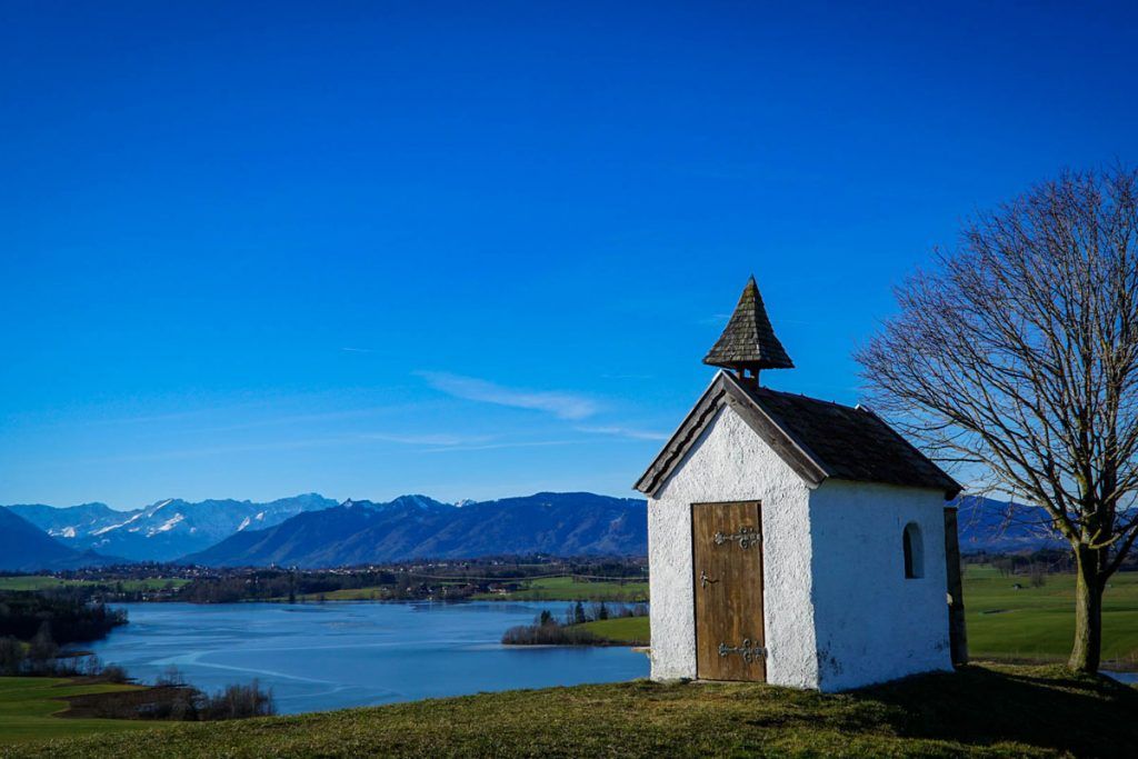 Die Mesnerhauskapelle Aidling im Hintergrund der Riegsee und Alpenpanorama