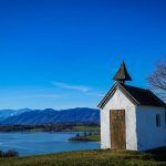 Die Mesnerhauskapelle Aidling im Hintergrund der Riegsee und Alpenpanorama